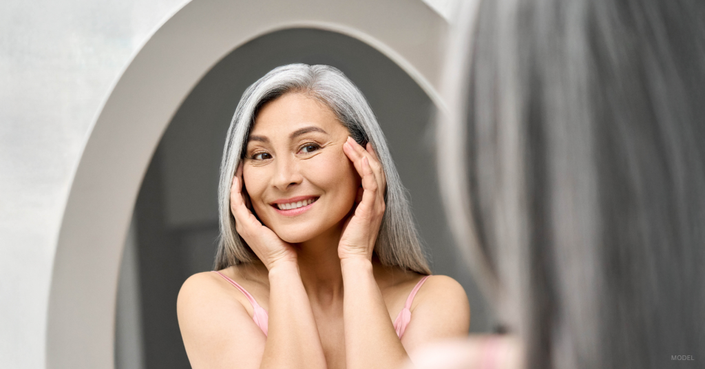 Woman looking in the mirror considering facelift vs liquid facelift. (Model)